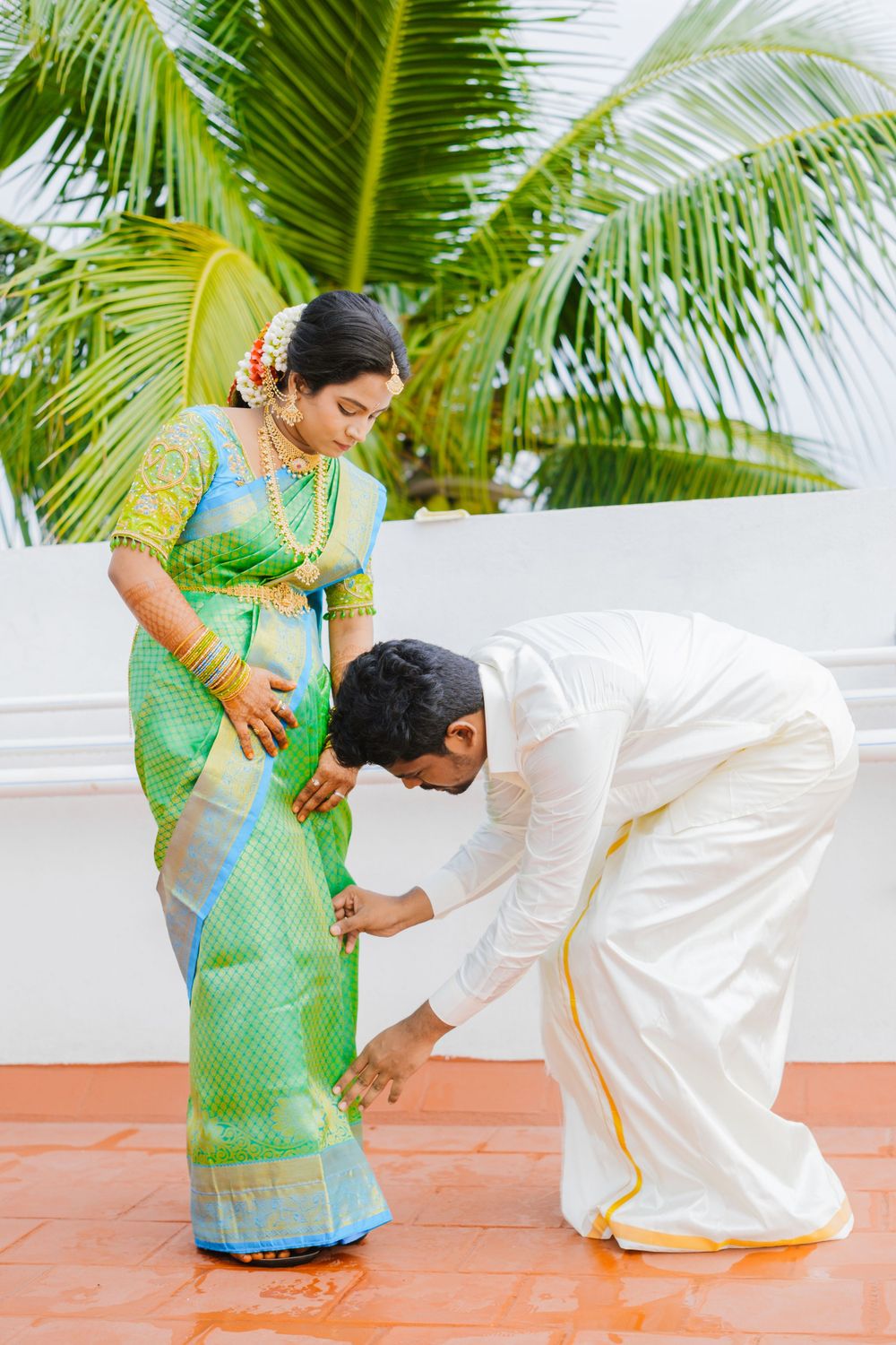 A traditional South Indian ceremony with a person in white attire bowing down to touch the feet of another in a green silk sari.