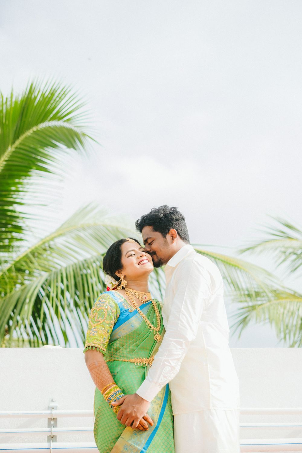 A romantic moment between a couple in traditional South Indian attire with palm trees in the background.