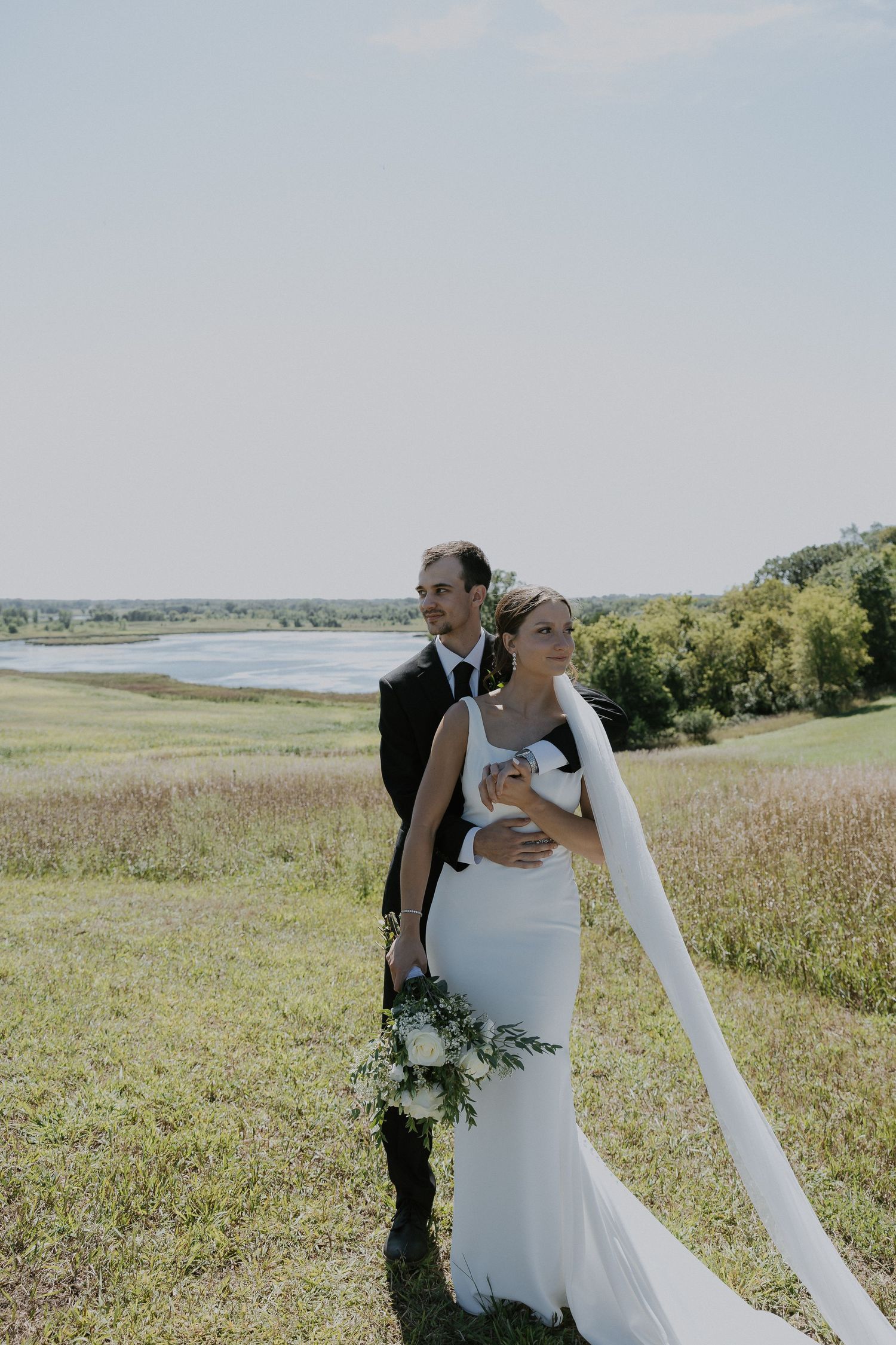Newlywed couple embraces on scenic hillside overlooking lake with flowing white wedding dress and floral bouquet.
