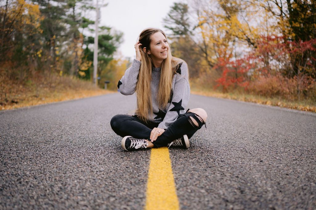 Person sitting cross-legged on yellow line of empty autumn road surrounded by fall foliage.