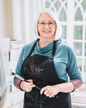 Artist in blue top and black apron poses with paintbrush in bright studio space.