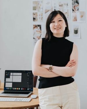 Professional in black sleeveless top stands confidently by desk with laptop and photo wall.
