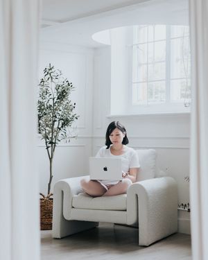 Minimal white room interior with person working on laptop in comfortable armchair near potted plant.