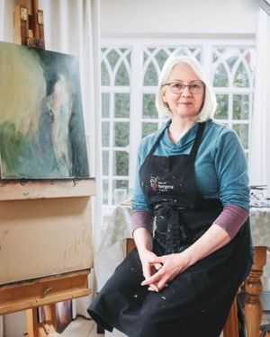 Artist wearing glasses and apron sits in studio with easel and artwork visible through arched windows.