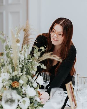 Someone in black arranges elegant floral decorations with wine glasses on a white table.