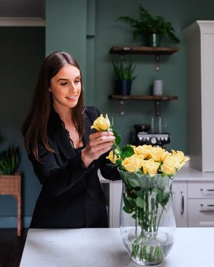 Woman arranging yellow roses in glass vase in modern kitchen with green walls.
