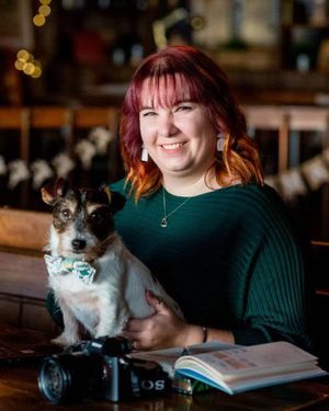 Person in green sweater poses with small black and white cat near camera and notebook.
