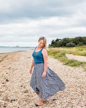 Person in blue tank top and floral skirt poses on beach with flowing fabric in wind.