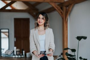 Professional wearing a light beige blazer and casual outfit stands in modern office space with exposed wooden beams.