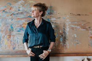 Someone in a navy blue button down shirt and dark jeans poses casually against a weathered wall with peeling paint.