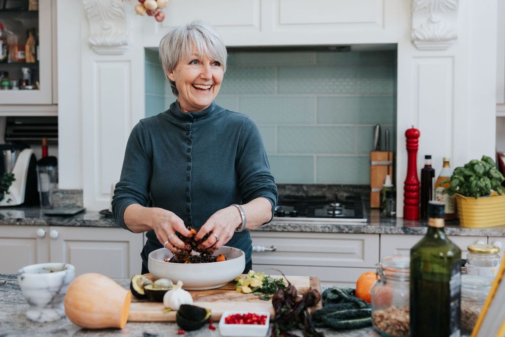 A person with silvery gray hair prepares food in a bright modern kitchen with marble countertops and light blue cabinetry.