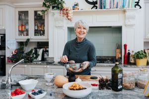 Someone in a teal turtleneck prepares food at a marble kitchen counter with fresh ingredients and cooking supplies spread out.