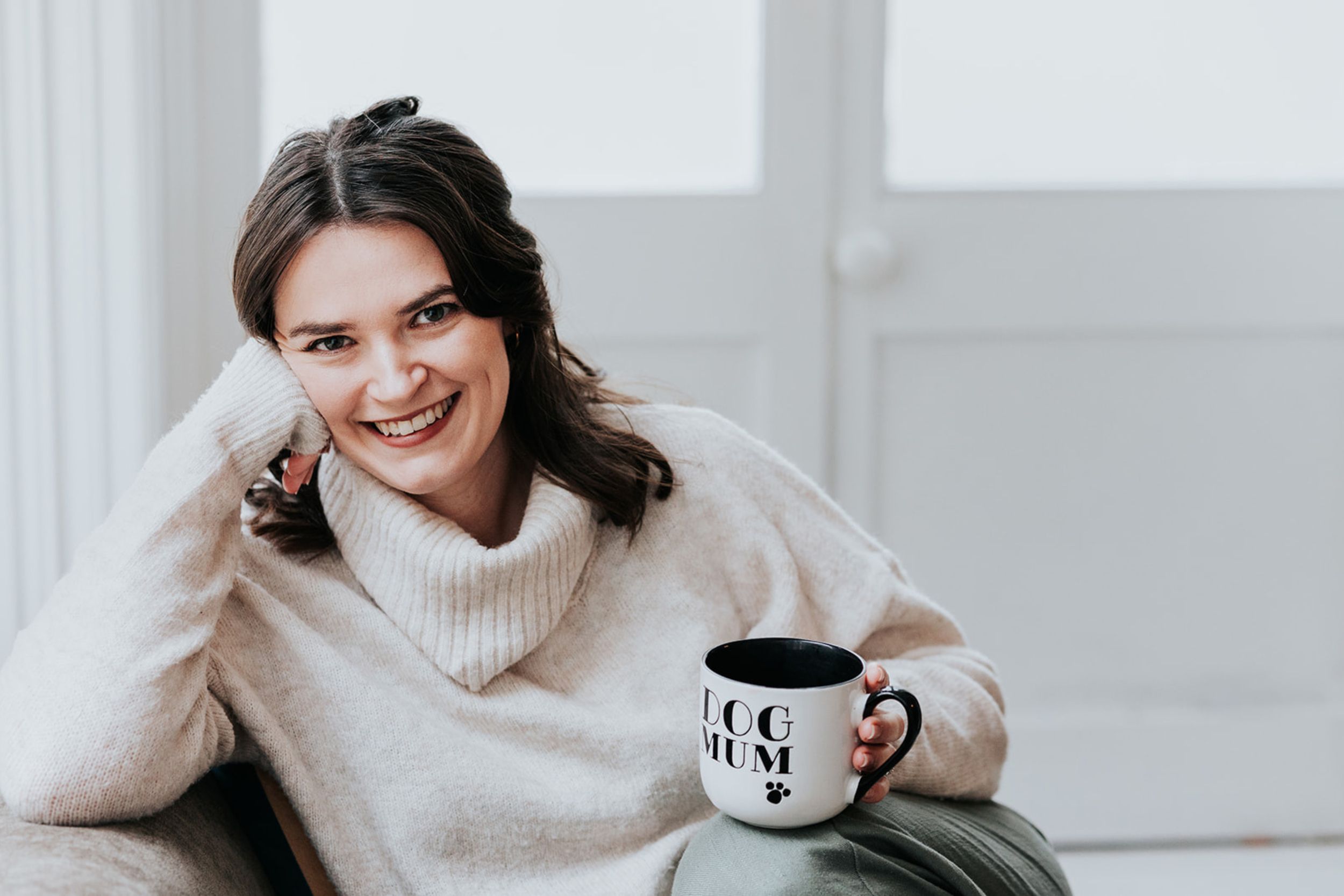 Someone in a cozy cream turtleneck sweater relaxes at home with a coffee mug and warm smile.