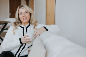 Someone in a white sweater with black trim sits comfortably on a couch while holding a coffee mug and smiling.