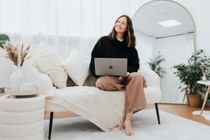 Remote worker in black sweater sits barefoot on modern white chair with MacBook laptop in minimal home office setting.