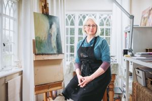 Artist wearing black apron stands in bright sunlit studio with paintings and art supplies.