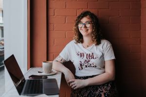Someone works at a cafe table with a laptop and coffee cup against an orange brick wall.