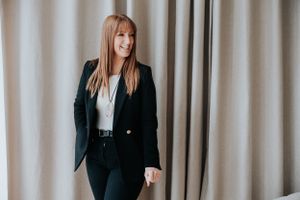 Professional woman in business attire poses in front of beige curtains wearing a black blazer and white blouse.