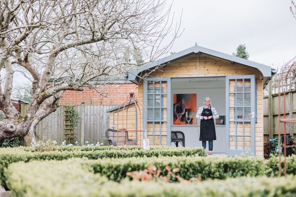 Two people stand at the doorway of a charming blue garden shed with boxwood hedges and bare tree branches overhead.