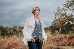 Person in white fluffy cardigan and patterned top poses outdoors against autumn landscape with dark stormy sky.