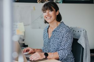 Professional wearing a patterned blouse sits at desk working on laptop with smartwatch and casual business attire.