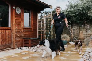 A professional dog handler stands with three dogs on leashes outside a wooden cabin on a stone patio.