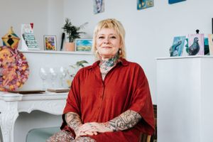 Someone in a red sweater with arm tattoos sits in a bright room decorated with colorful artwork on the walls.