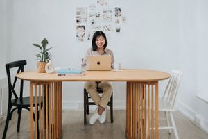 A person works on a laptop at a wooden dining table with indoor plants and wall decor in a bright, minimalist space.