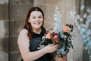 Smiling person in black sleeveless dress holds beautiful bouquet of peach roses and delicate blue flowers against neutral wall.