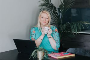 Professional woman in turquoise outfit sits at desk with laptop and notebook in modern workspace.
