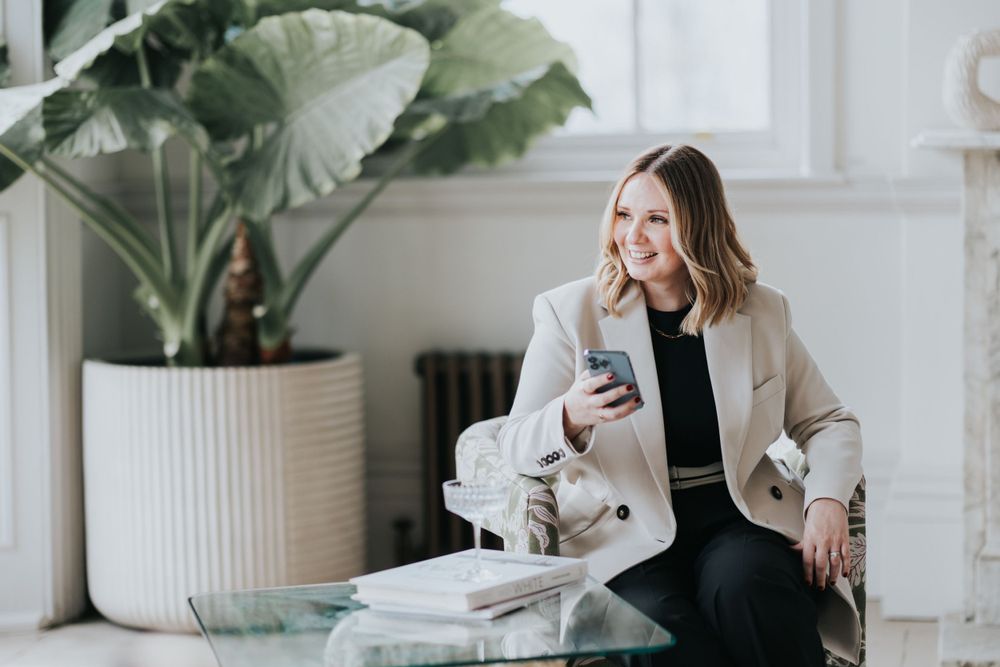 Professional woman in beige blazer sits by potted plant while working on smartphone in bright modern space.