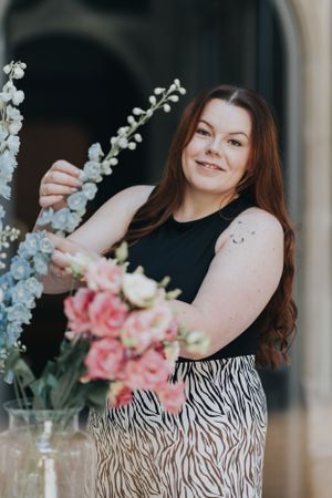 A person in a black top and zebra print skirt arranges white delphinium and pink roses in a glass vase.