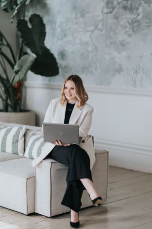 Professional sits on grey couch with laptop in bright modern room with houseplant and textured wall.