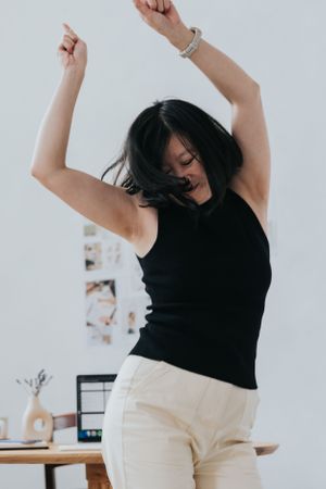 Dancing figure in black sleeveless top and white pants moves energetically against white wall with desk in background.