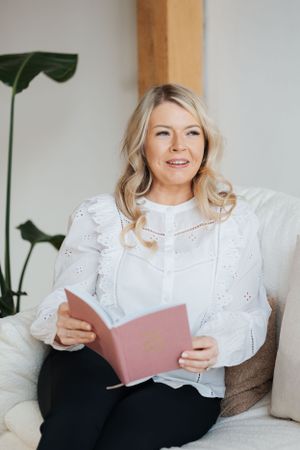 Someone in a white eyelet lace blouse sits comfortably on a couch reading a pink book near a large houseplant.