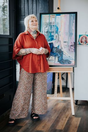 Artist wearing a red blouse and floral pants stands smiling next to an easel with artwork in a cozy studio space.