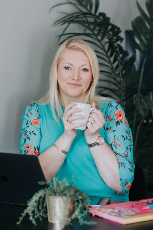 Person in turquoise floral top enjoying coffee at desk with plants and books in cozy indoor setting.