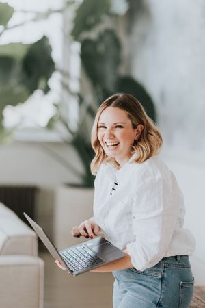A person in a white blouse and jeans stands smiling while holding a laptop in a bright, modern space.