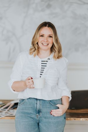 A person in a white button-down shirt and striped tee holds a coffee cup while standing against a gray wall.