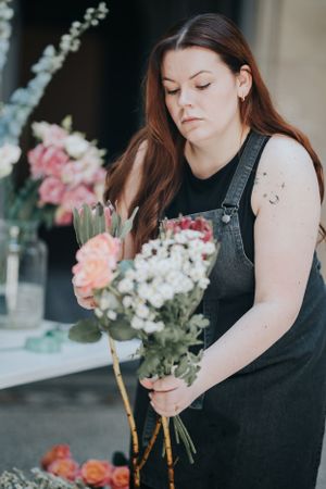 A florist in denim overalls arranges fresh pink and white flowers in a creative composition near a window.