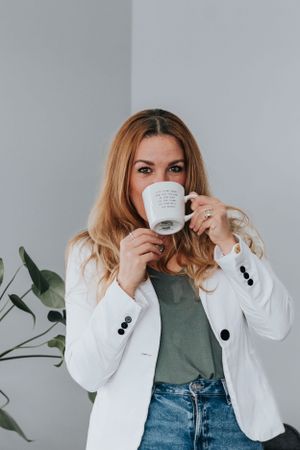Woman in white blazer and jeans holds white coffee mug while standing against light gray wall with plant decor.