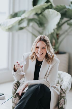 Candid lifestyle shot of someone in a cream blazer enjoying a cocktail while relaxing on a couch with plants in background.