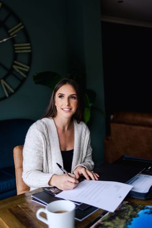 A person sits at a wooden table with paperwork, a coffee cup, and a large clock visible on the teal wall behind them.