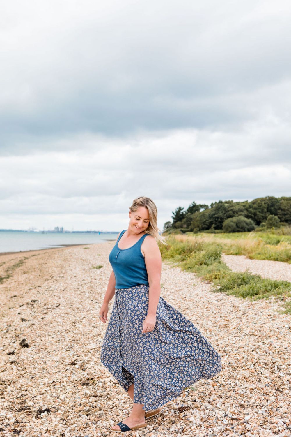 Woman in blue tank top and flowy floral skirt poses on a pebble beach with trees and cloudy sky in background.