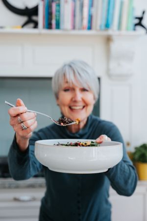 Person in blue sweater holding white bowl with food, smiling while preparing to eat a meal in bright kitchen setting.