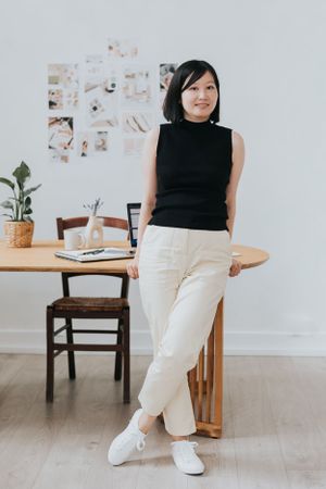 Casual business style with black sleeveless top and white pants, leaning against wooden table in minimalist home office space.