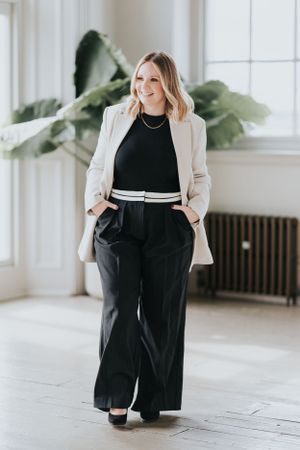 A person wearing a beige blazer, black top, and wide-leg black pants poses confidently in a bright interior space with plants.