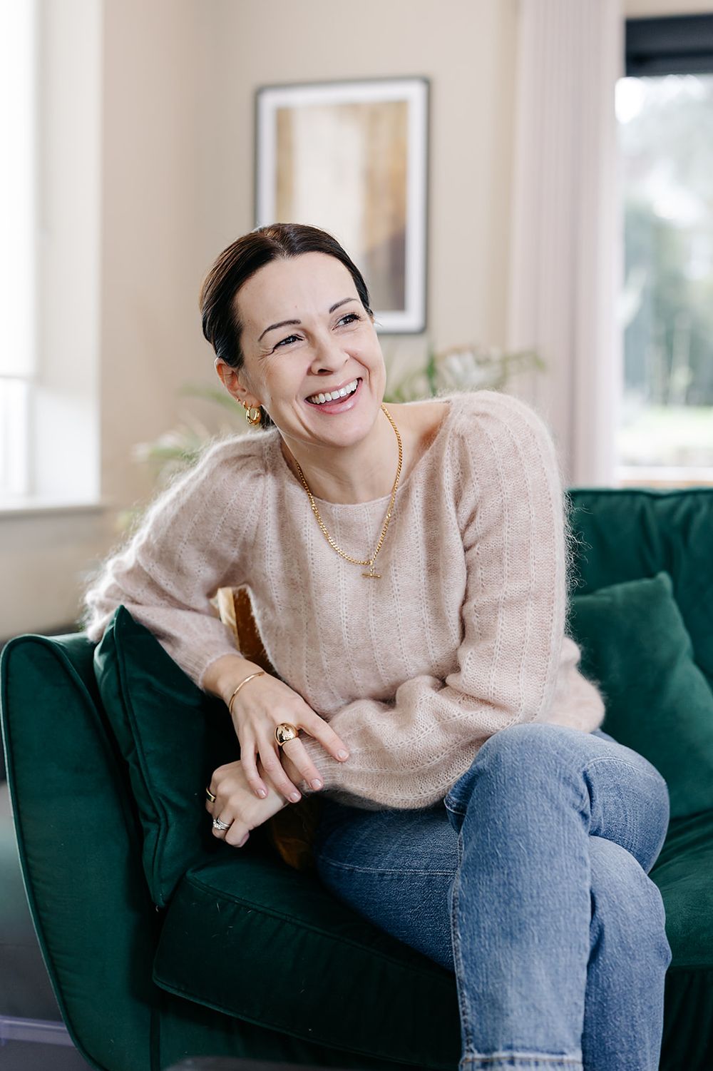Someone relaxes comfortably on a dark green velvet couch wearing a beige sweater and blue jeans in a bright room.