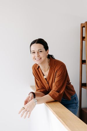 Person in brown shirt leaning on wooden table and smiling in minimalist interior setting.