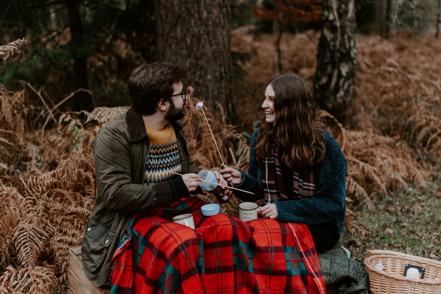A couple are sat in the woods, toasting marshmallows and making hot chocolate drinks.